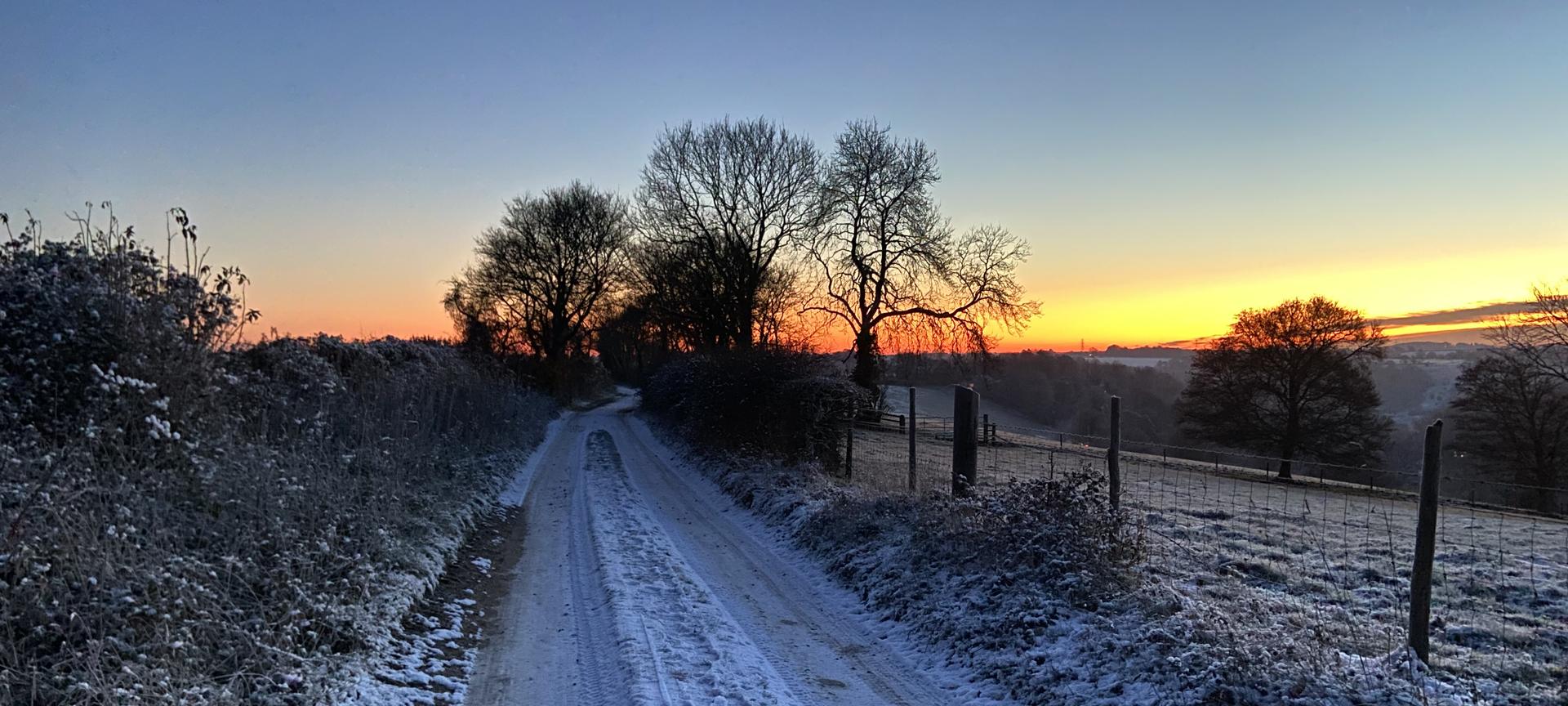 Frosty farm track at dawn, with bare trees silhouetted against an orange sky