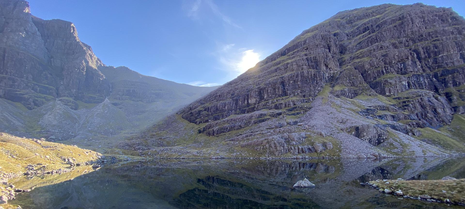 Sun breaking over the edge of a steep mountain corrie above a still lochan