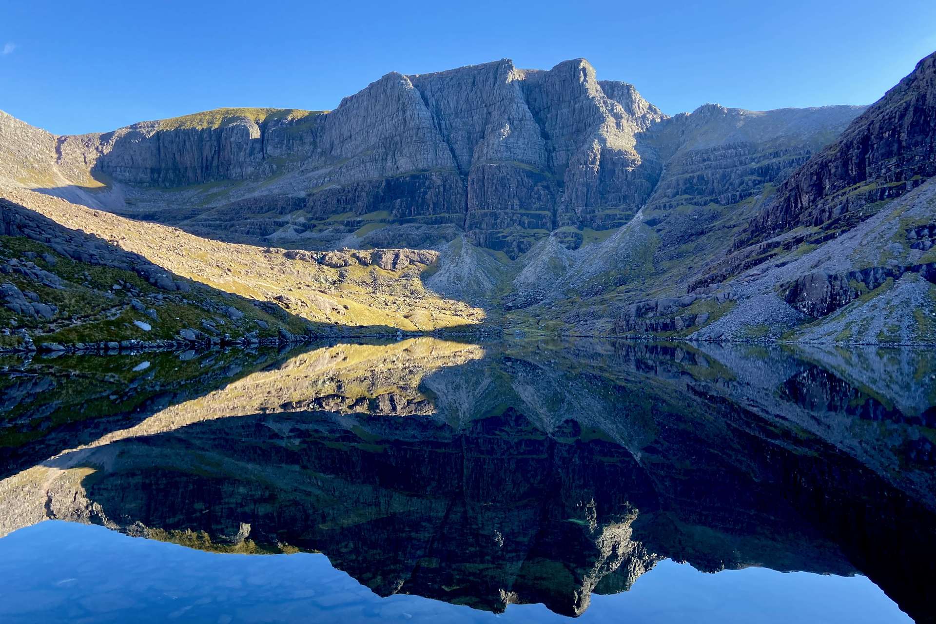 Still mountain loch reflecting towering sandstone cliffs under a clear blue sky