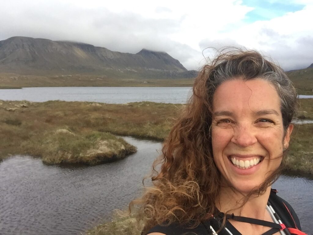 Louise Watson smiling beside a Highland lochan, with mountains behind